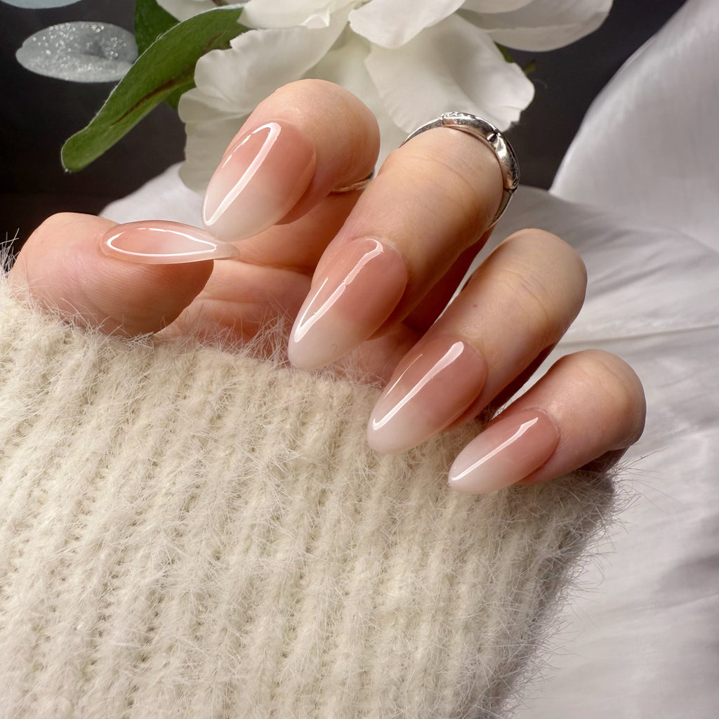 A hand wearing short almond-shaped press-on nails with a glossy nude-to-white ombre gradient, paired with a silver ring and a white fuzzy sweater.