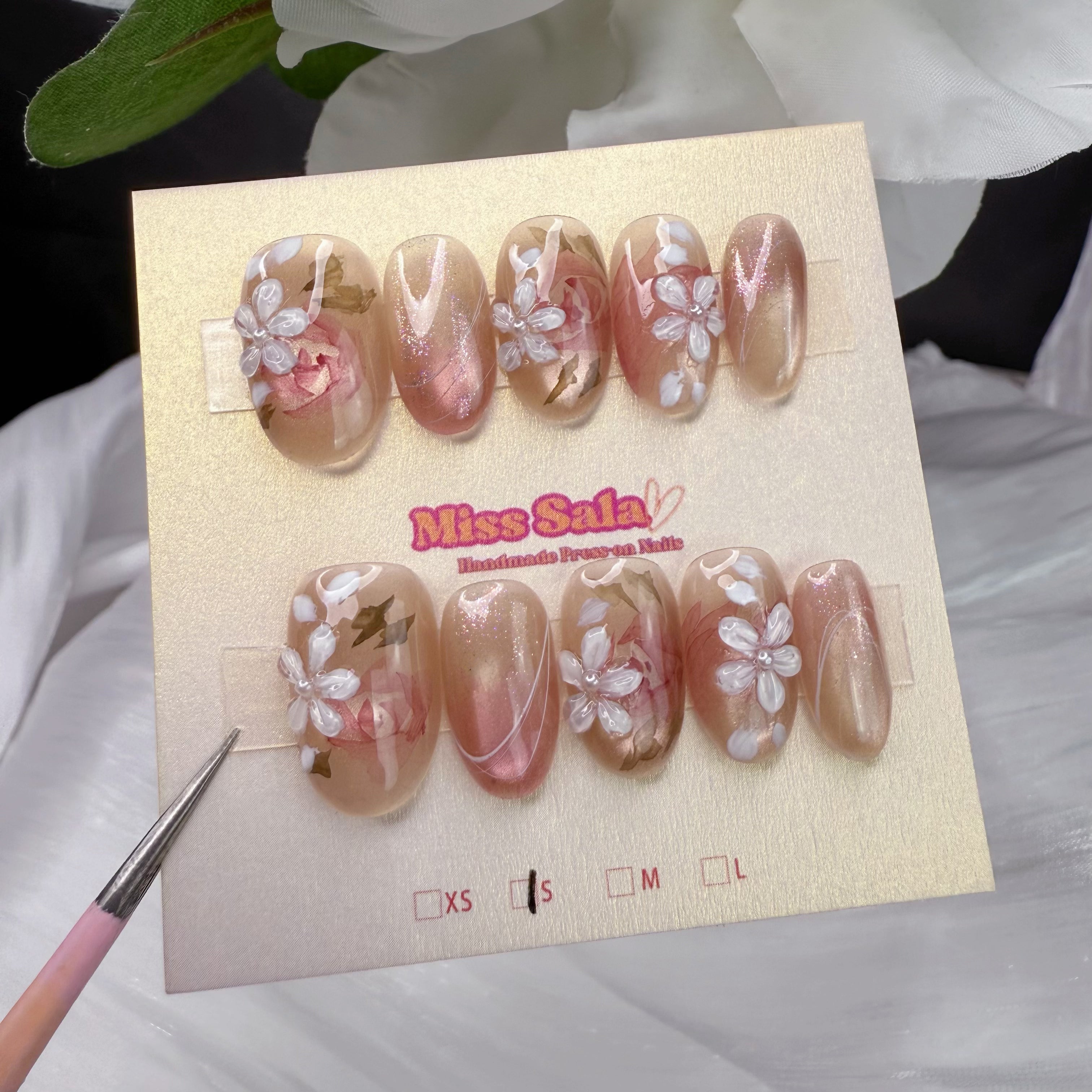 Almond-shaped press-on nails featuring a shimmering nude-pink base, decorated with delicate 3D white flower charms, hand-painted green leaf details, and subtle pearl accents.