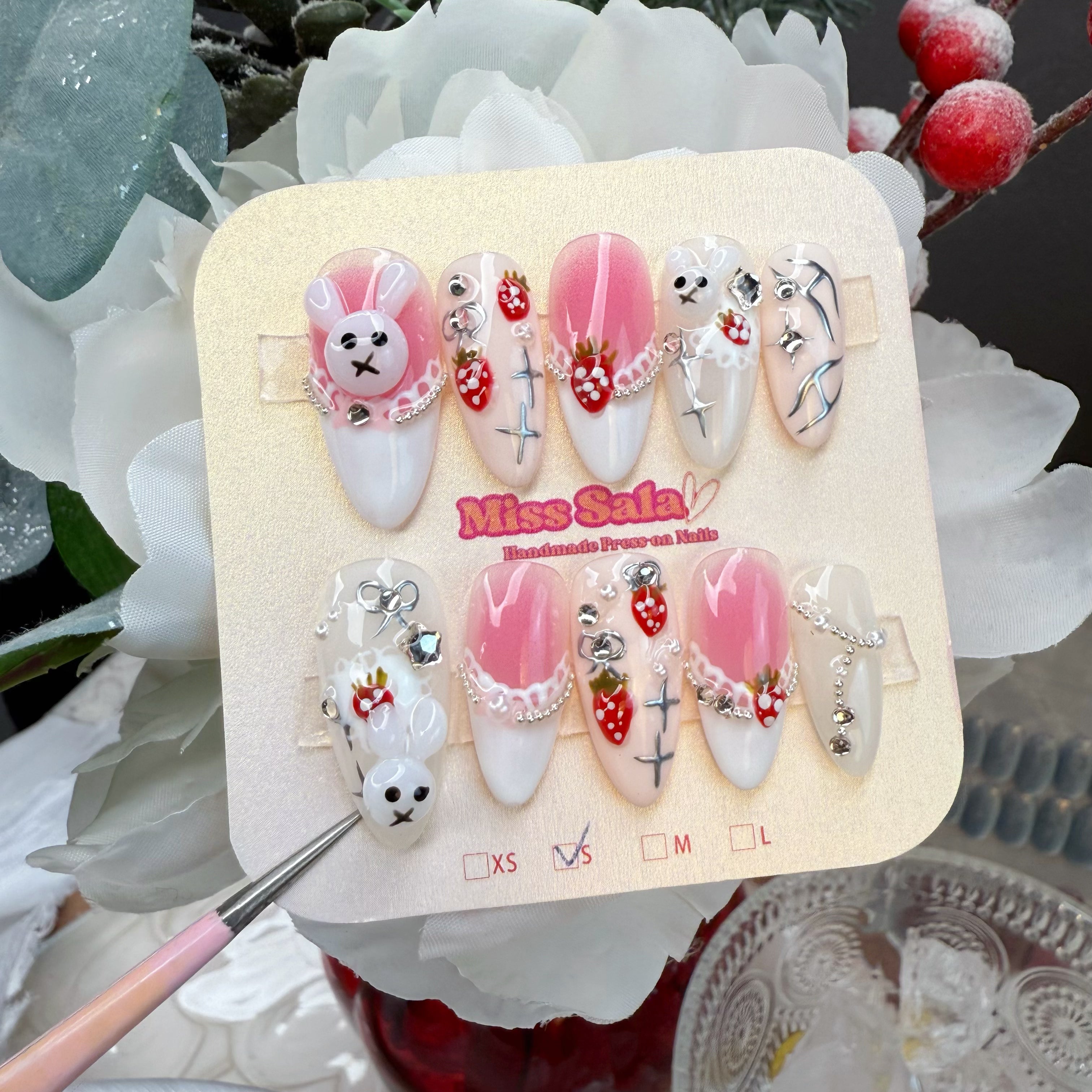 Almond-shaped press-on nails featuring 3D bunny charms, hand-painted strawberries, delicate lace detailing, and silver chain accents on a pink and white base.
