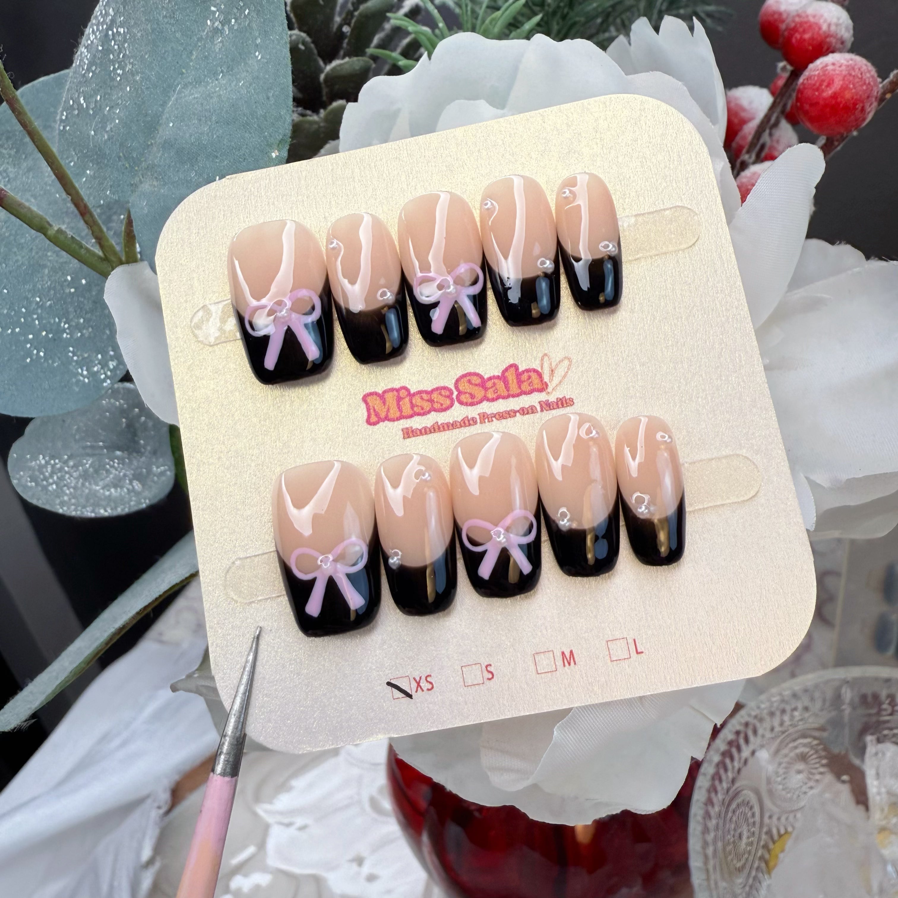 Short square press-on nails featuring a glossy nude base with black French tips, adorned with hand-painted pink bows and delicate silver pearl accents.