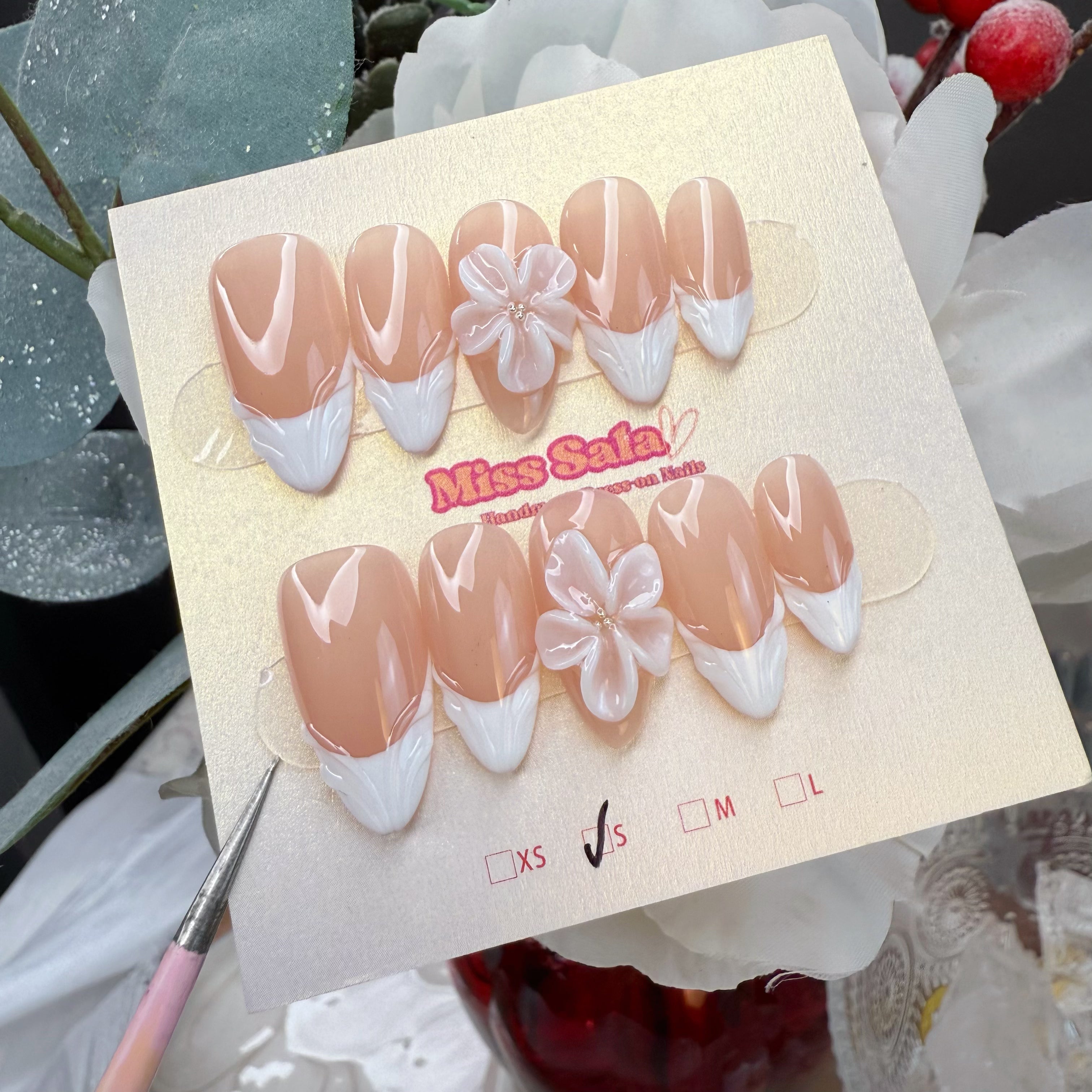 Almond-shaped press-on nails with a glossy nude base, featuring white 3D textured French tips and large iridescent 3D flower charms with pearl centers.
