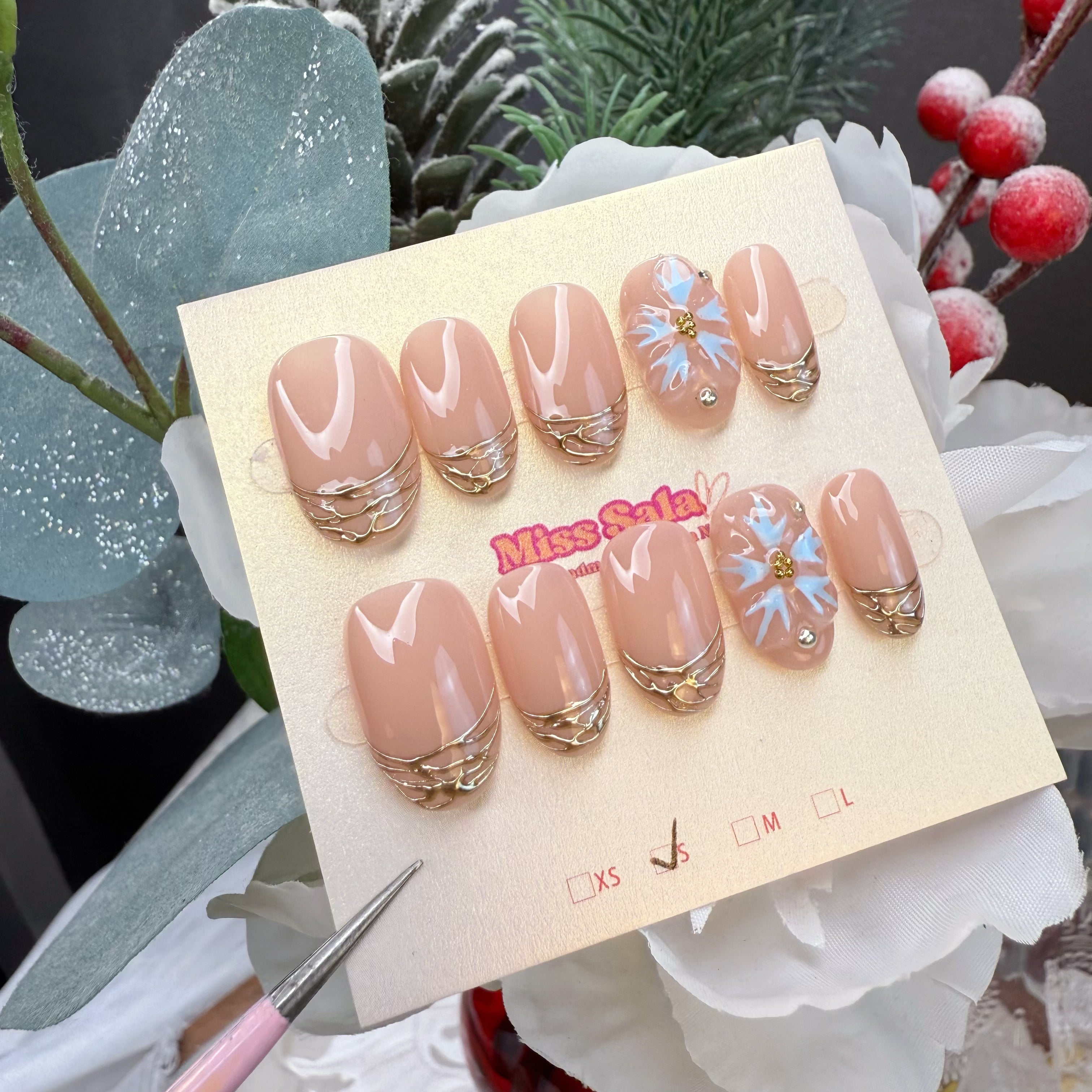 Almond-shaped press-on nails featuring a glossy nude base, hand-applied gold chrome wire-effect French tips, and 3D light blue flower charms with crystal centers.
