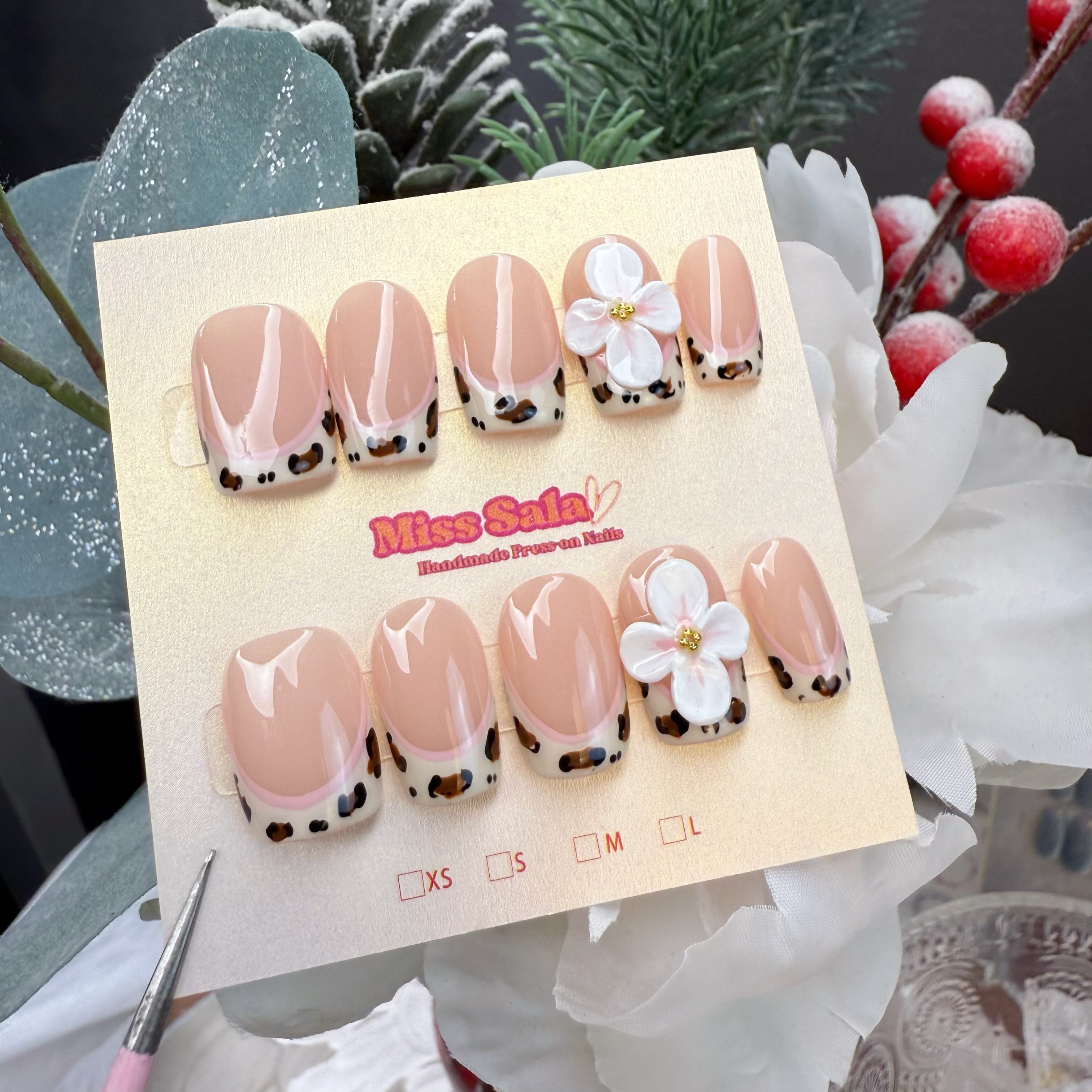 Short square press-on nails featuring a nude base with leopard print French tips, pink line art, and large 3D white flower charms with gold centers.