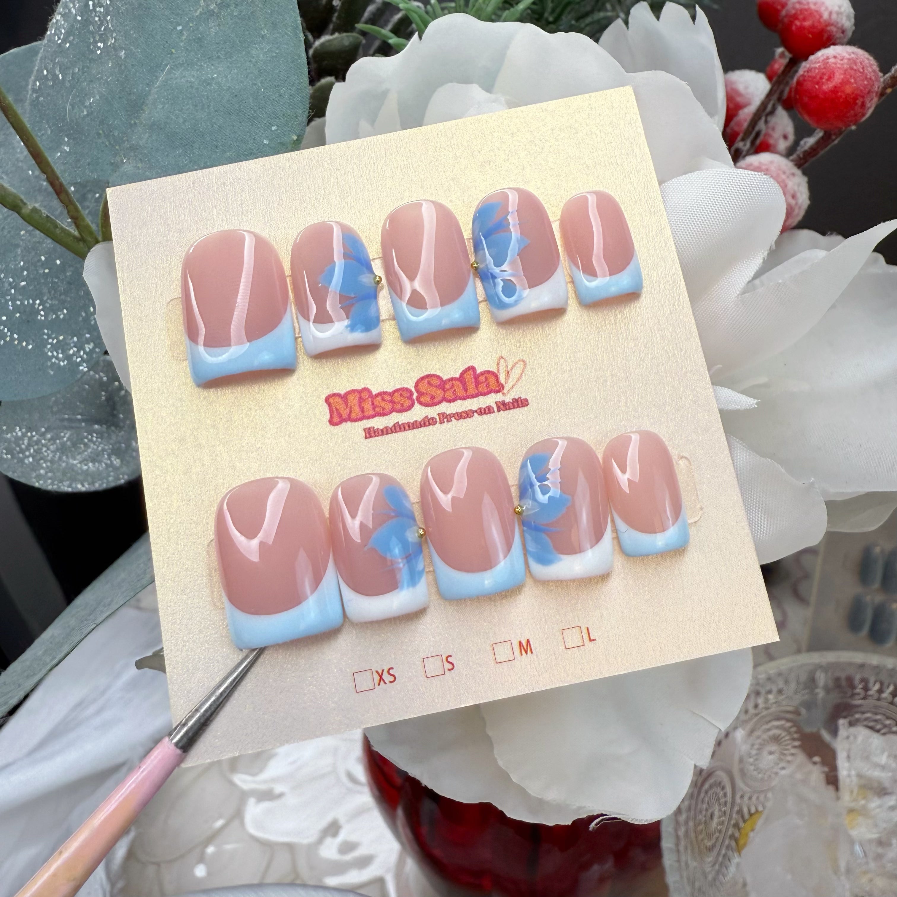 Short square press-on nails featuring a glossy nude base, sky-blue French tips, and delicate hand-painted blue ribbons with tiny gold bead centers.