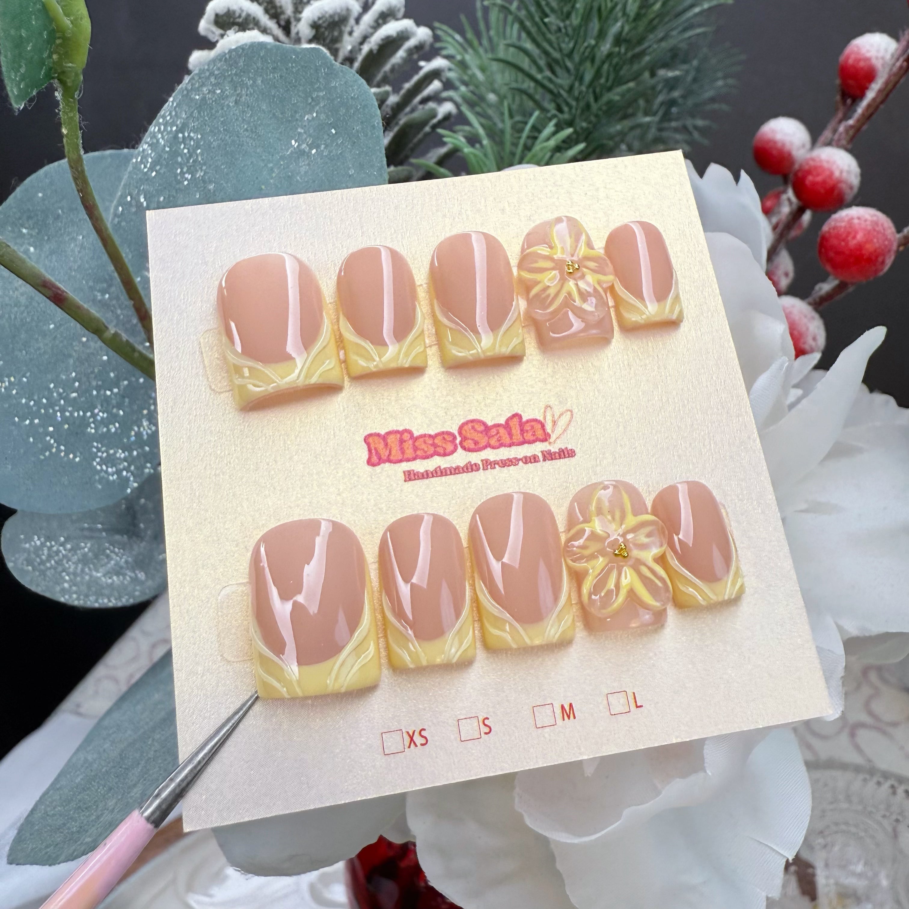 Short square press-on nails with a glossy nude base and yellow petal-style French tips, featuring large 3D yellow flower charms with gold center accents.
