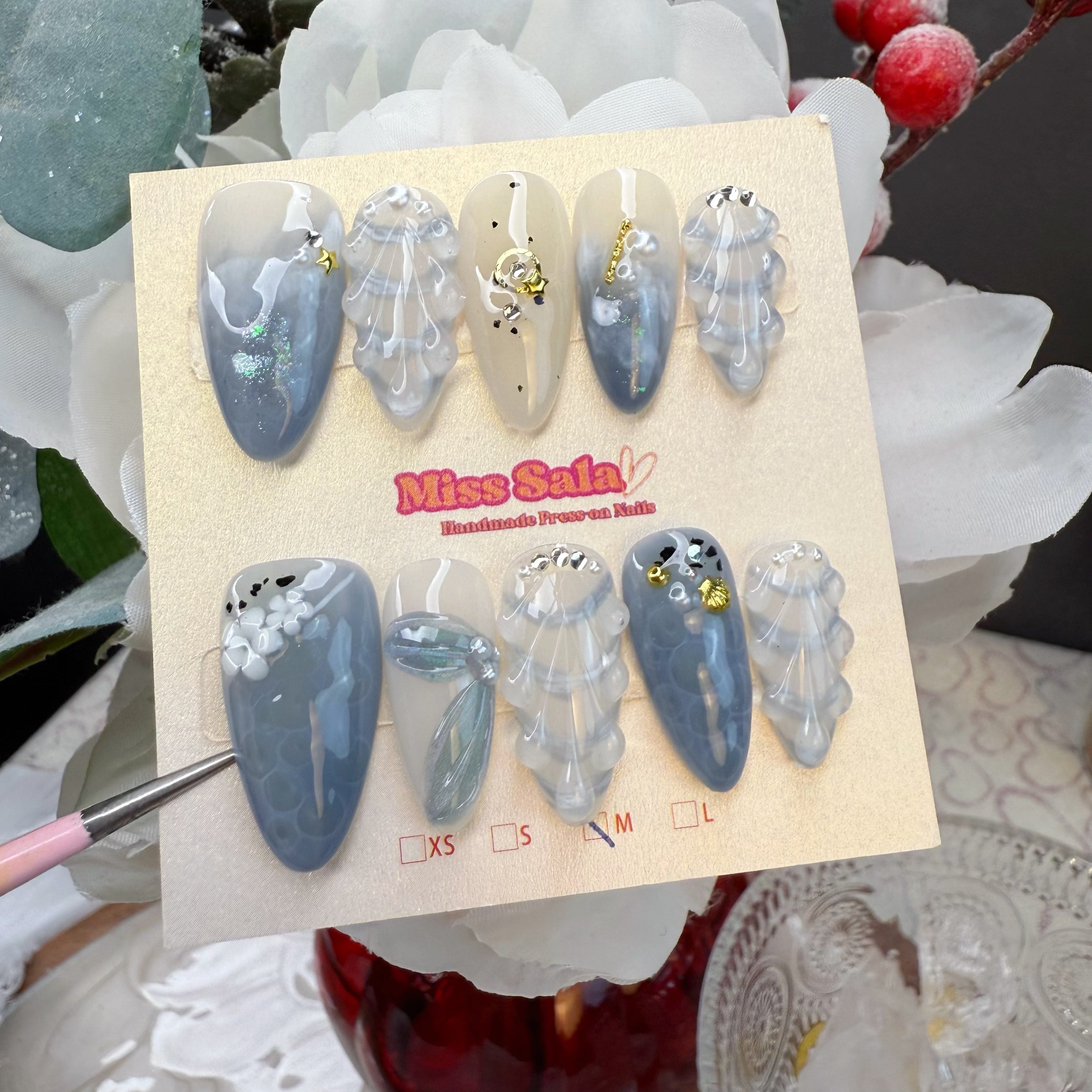 Almond-shaped press-on nails featuring a blue and white marble design, 3D textured shell art, and celestial gold charms with crystal accents.