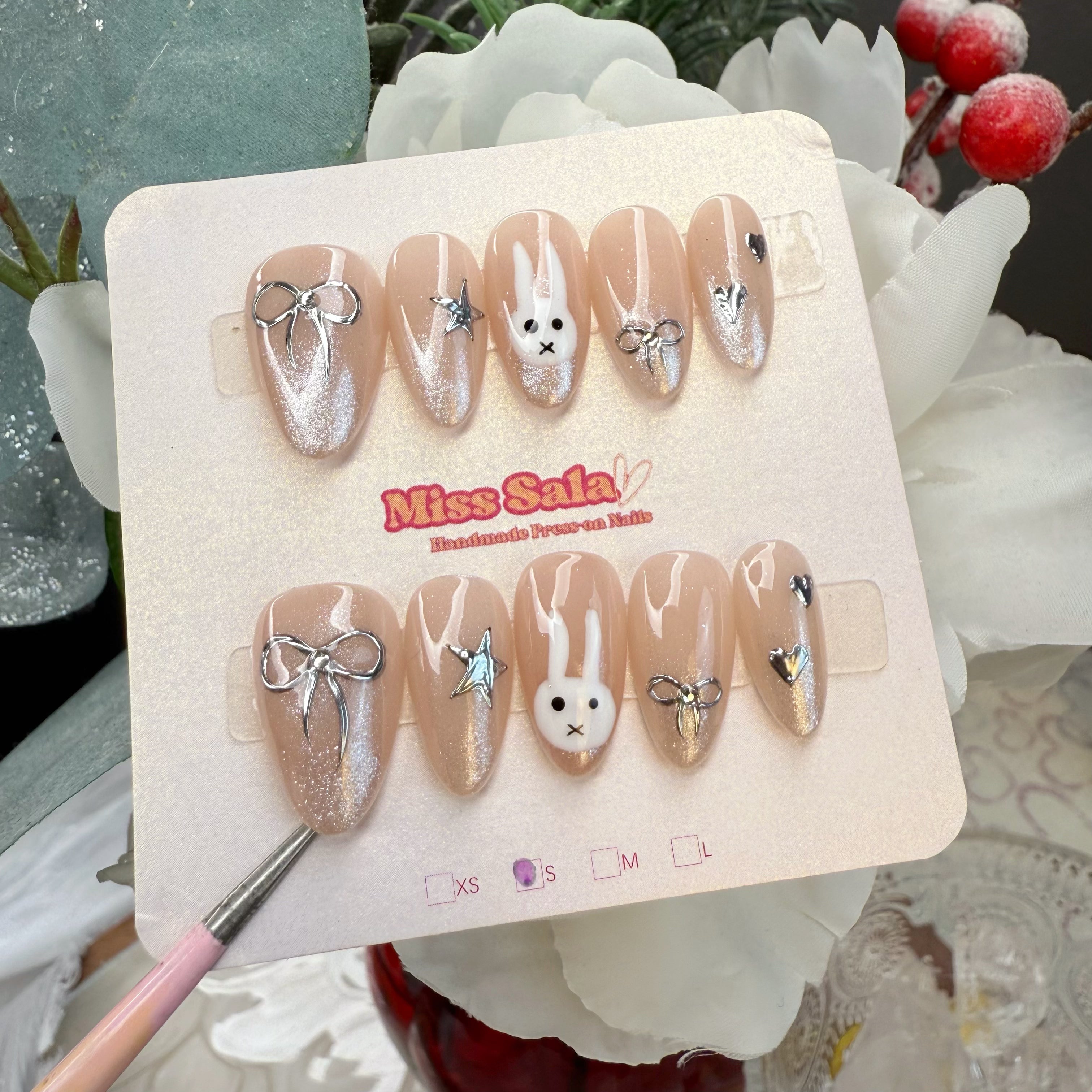 Almond-shaped press-on nails with a nude cat-eye magnetic shimmer base, featuring a hand-painted white bunny face, 3D silver chrome bows, hearts, and stars.
