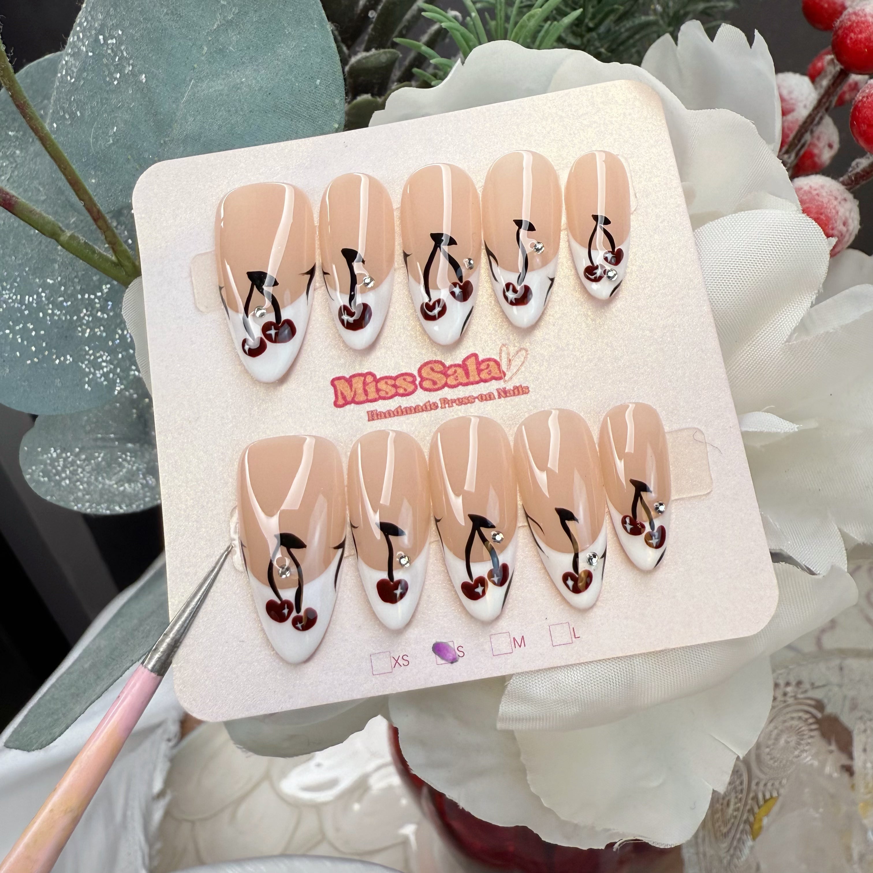 Almond-shaped press-on nails featuring a white French tip base with hand-painted dark red cherries, black stems, and tiny crystal rhinestone accents.