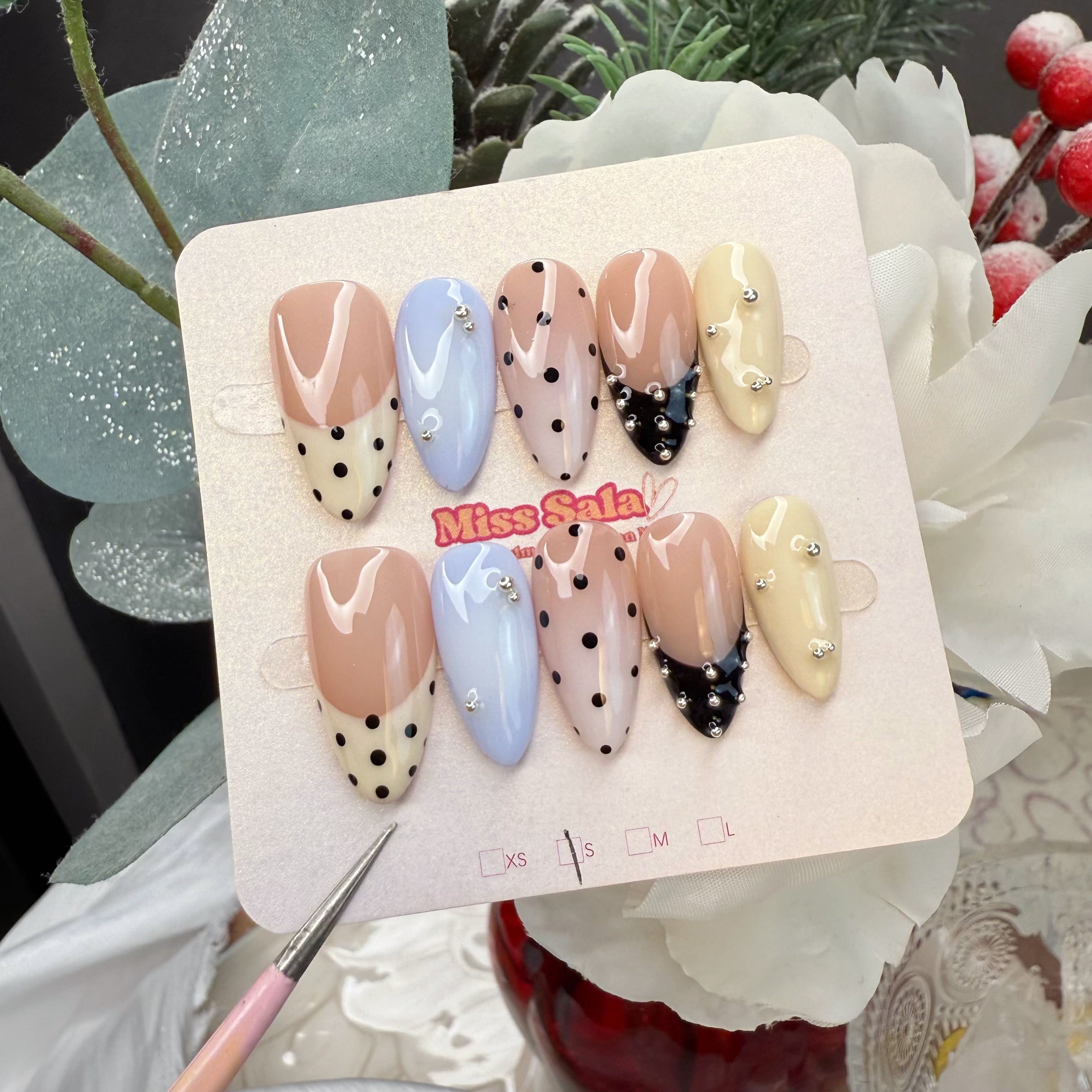 Almond-shaped press-on nails featuring a mix of nude, pastel blue, and cream bases with black polka dots, black French tips, and silver bead accents.