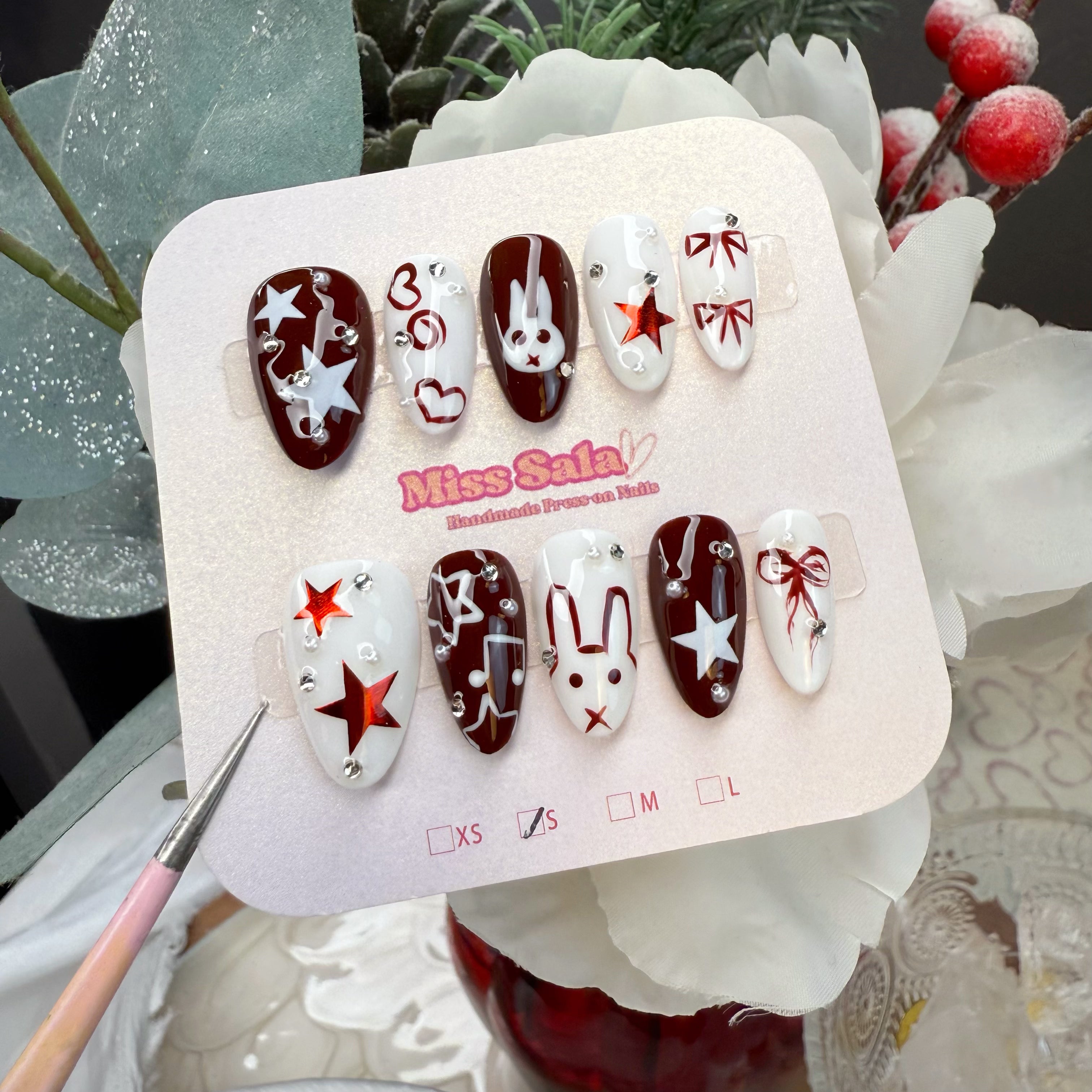 Almond-shaped press-on nails in a high-contrast deep red/ black and white theme, featuring hand-painted bunny faces, metallic stars, and delicate red bows with crystals.
