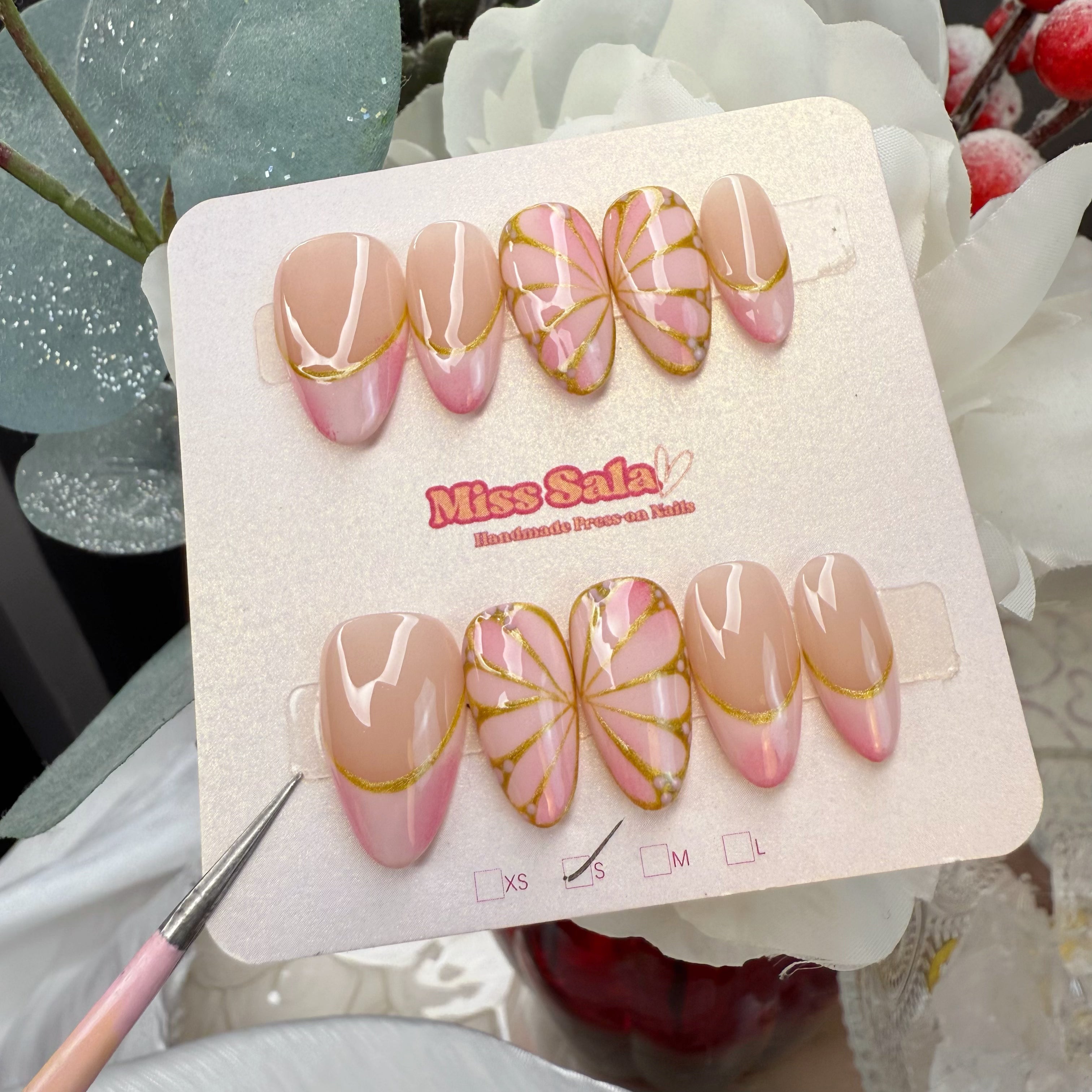 Almond-shaped press-on nails featuring soft pink French tips with gold glitter outlines and detailed hand-painted pink and gold butterfly wing art.