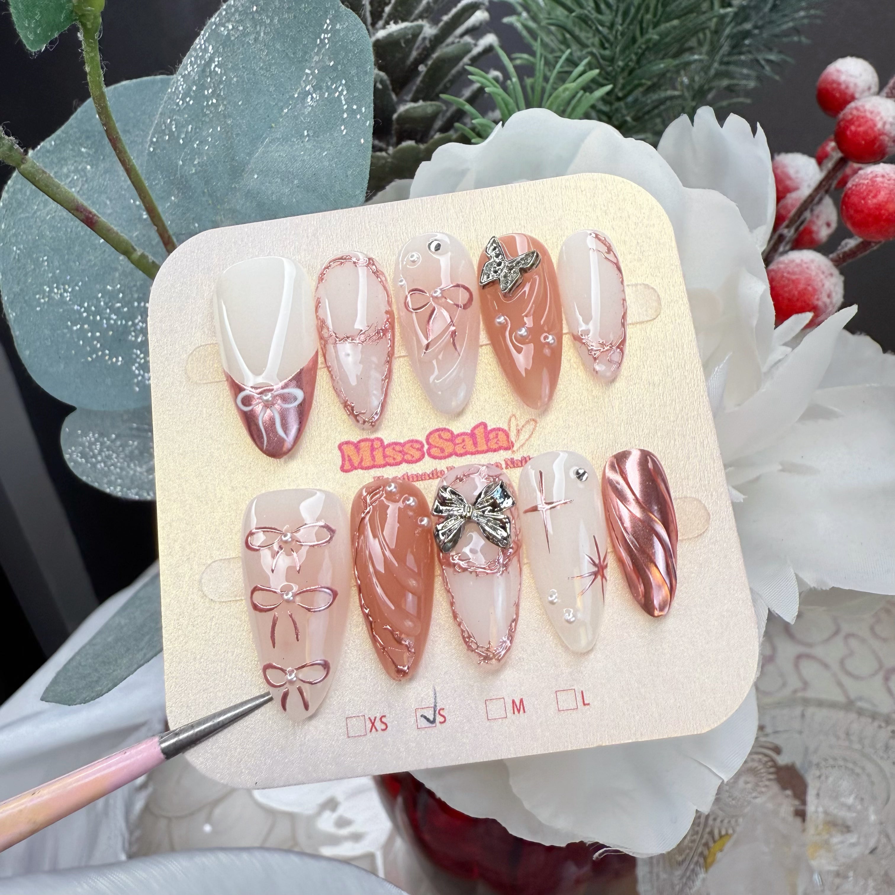 Almond-shaped press-on nails with a nude and white base, featuring rose gold metallic 3D bows, butterfly charms, textured rose gold borders, and shimmering star accents.