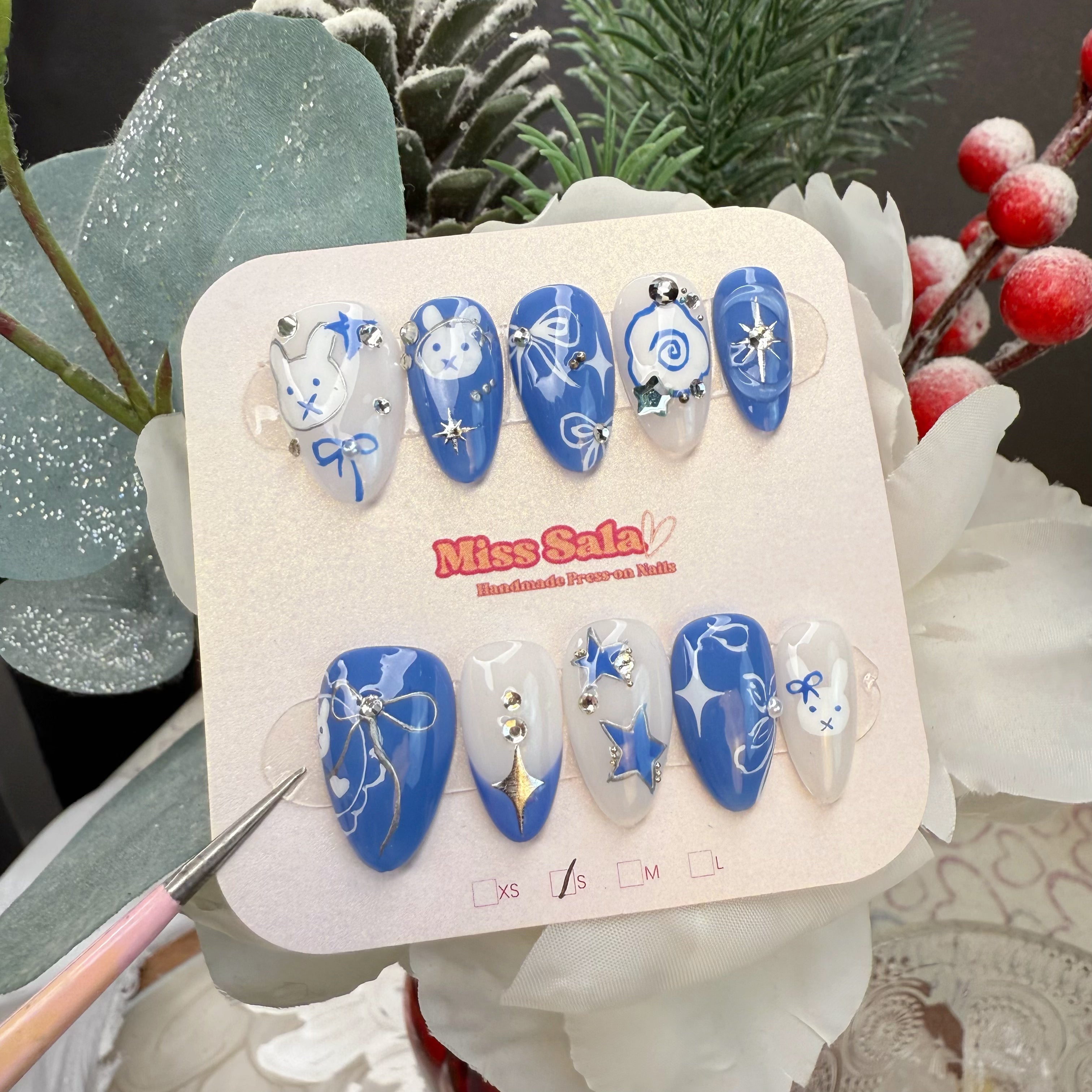 Almond-Shaped Press-On Nails in Blue and White with delicate raised Bunny Accents, Silver Metallic Stars, Blue Ribbons, and Sparkling Rhinestones.