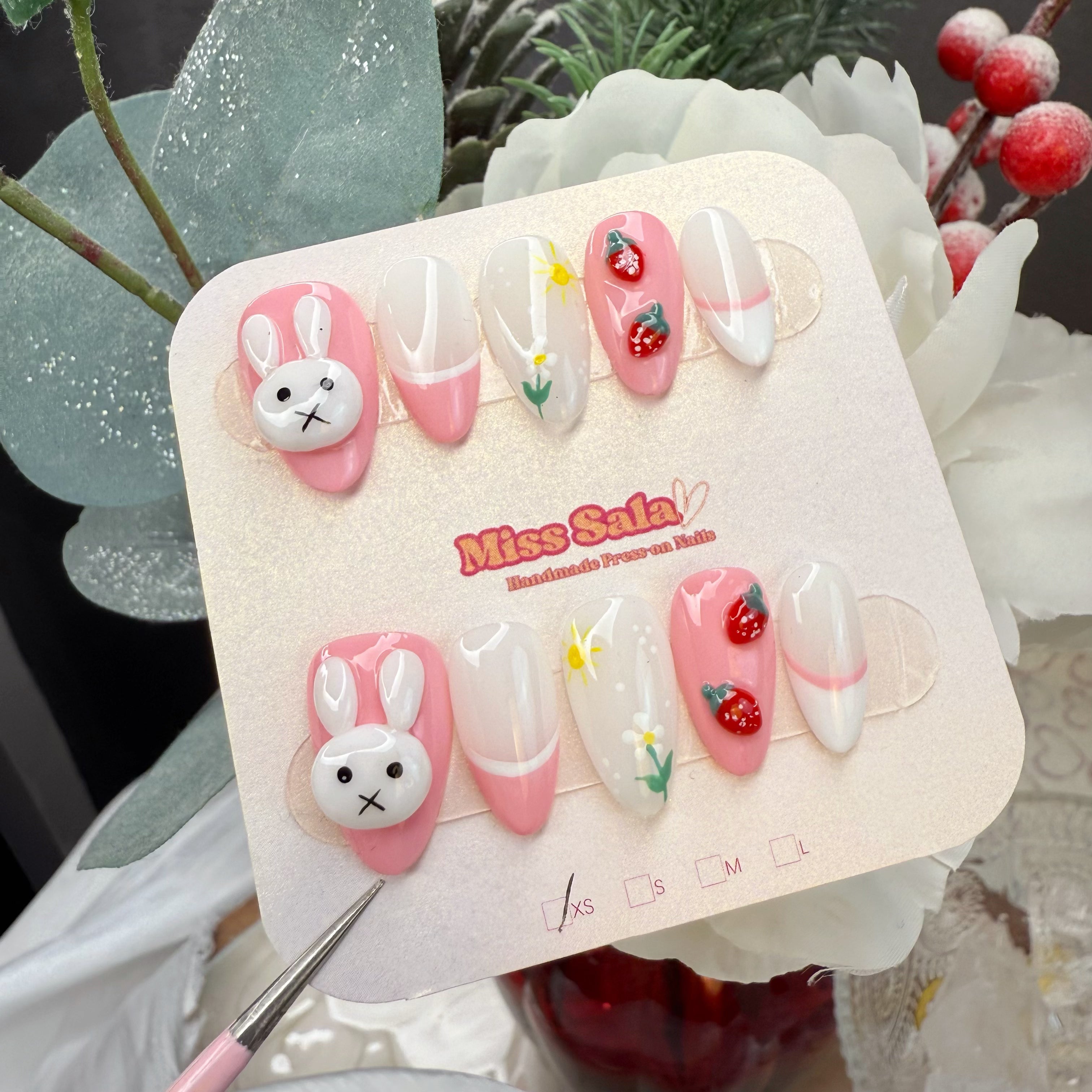 Almond-shaped press-on nails with a pink and white base, featuring a large 3D white bunny charm, 3D red strawberries, and hand-painted daisies and sun motifs.