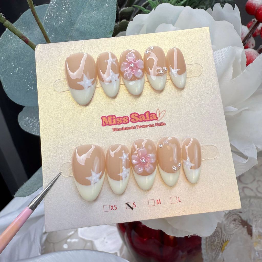 Almond-shaped press-on nails with a nude base and cream French tips, featuring a 3D pink resin flower with a pearl center, hand-painted white starbursts, and tiny pearls.