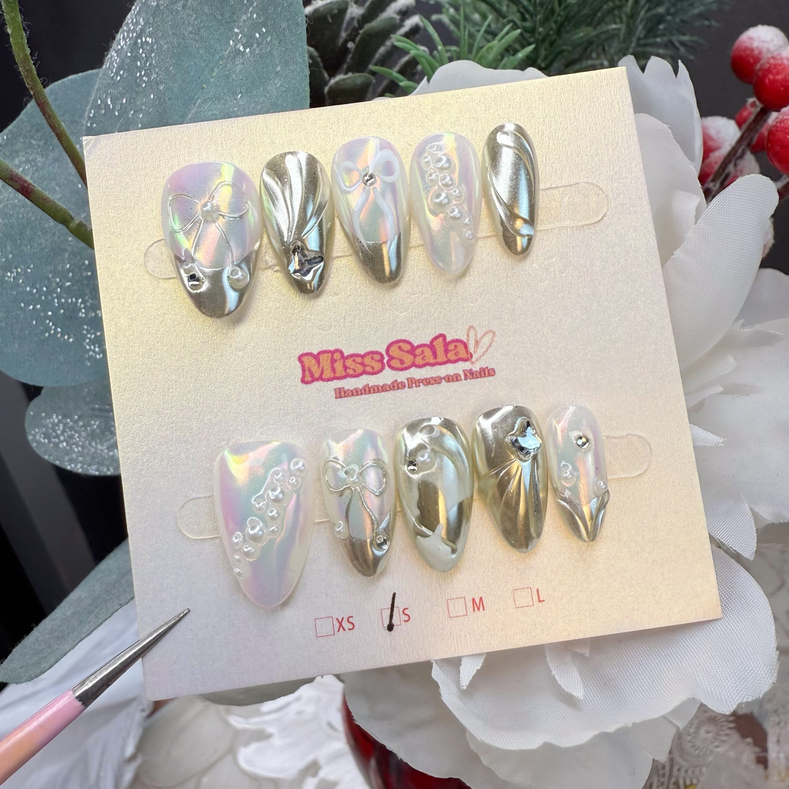 Almond-shaped press-on nails featuring a high-shine silver liquid chrome finish, 3D iridescent pearl accents, sculpted silver bows, and metallic wave textures.