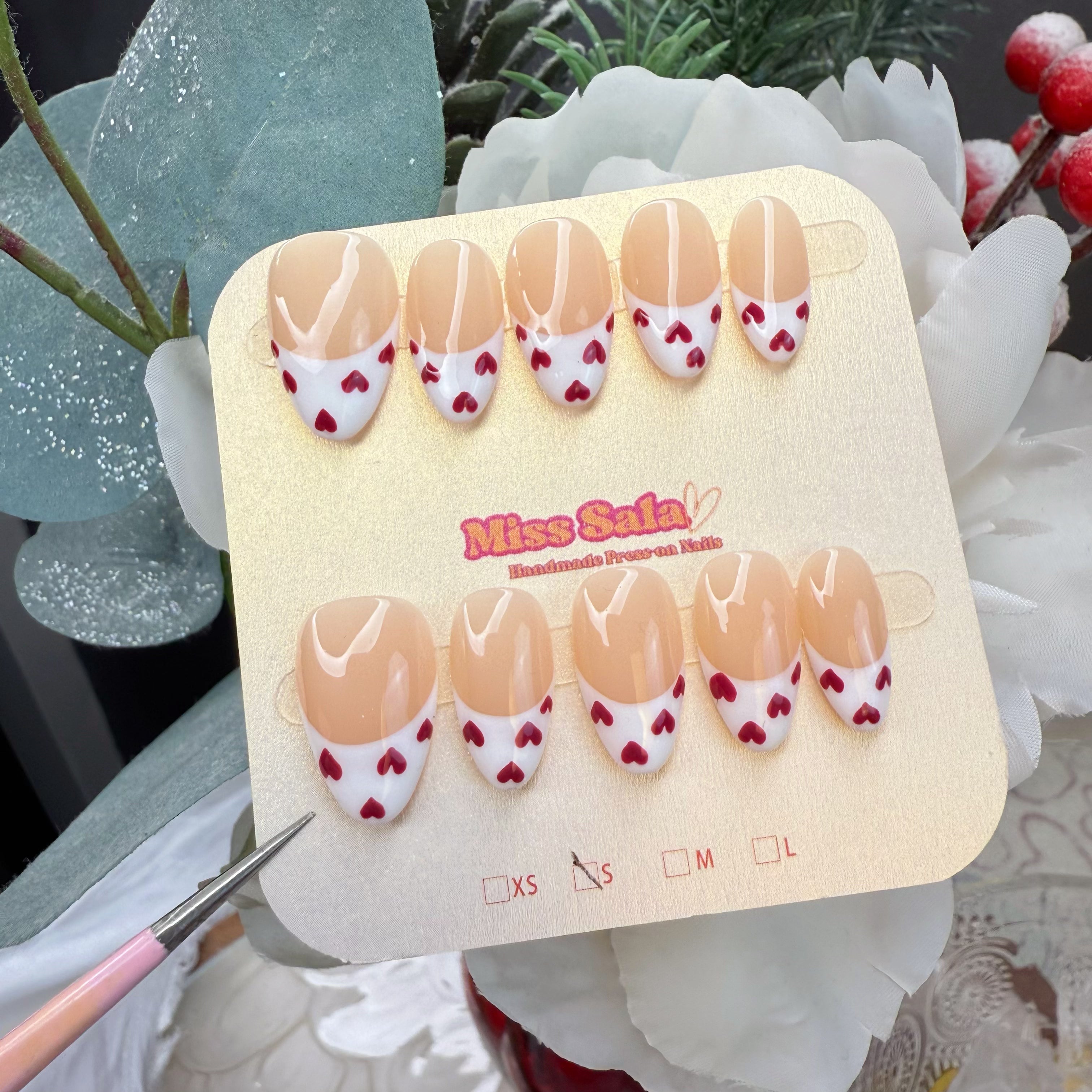 Almond-shaped press-on nails featuring a nude base with crisp white French tips and a delicate pattern of small hand-painted red hearts.