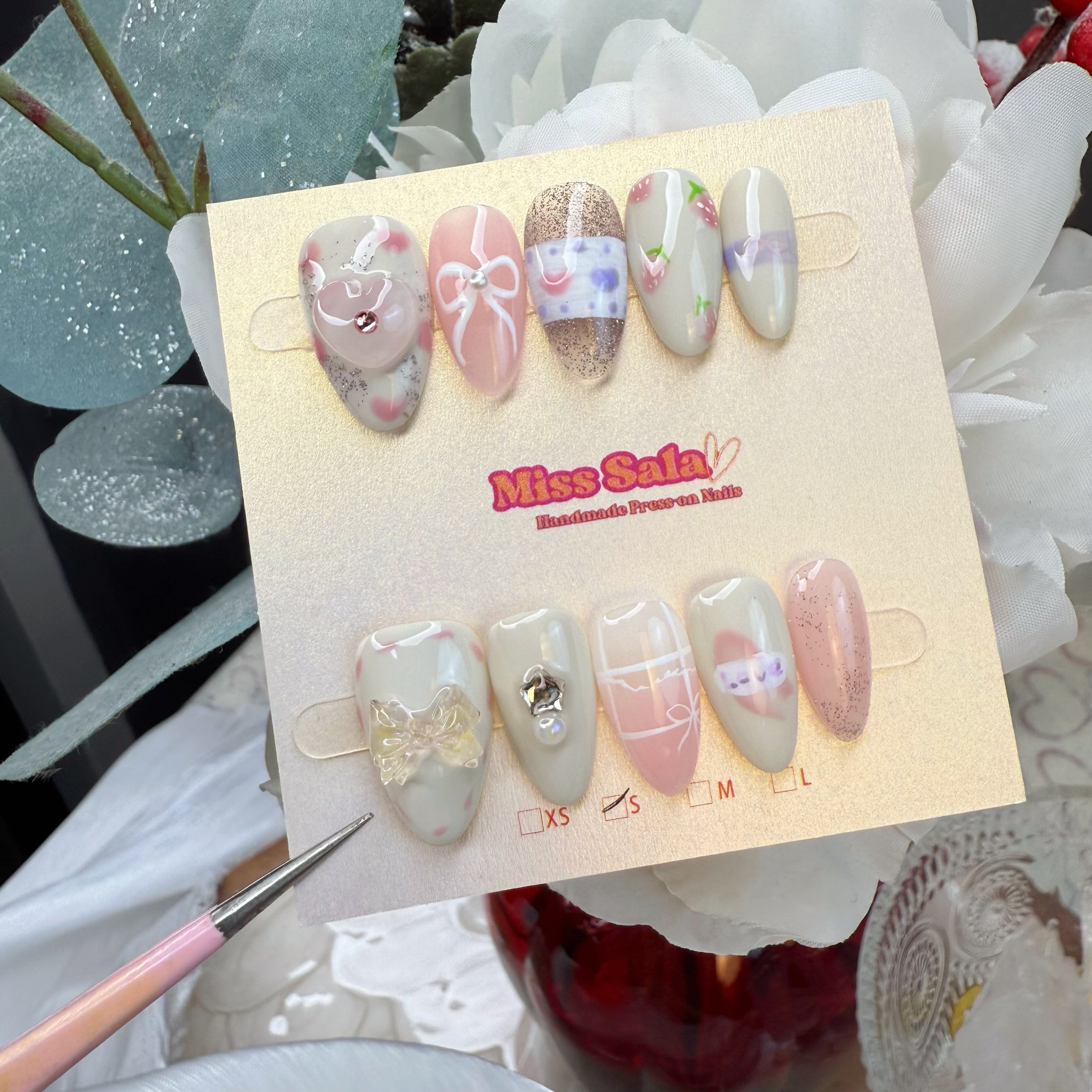 Almond-shaped press-on nails featuring hand-painted pink bows, delicate heart motifs, 3D heart and bow charms, and silver glitter accents on a pastel pink and cream base.