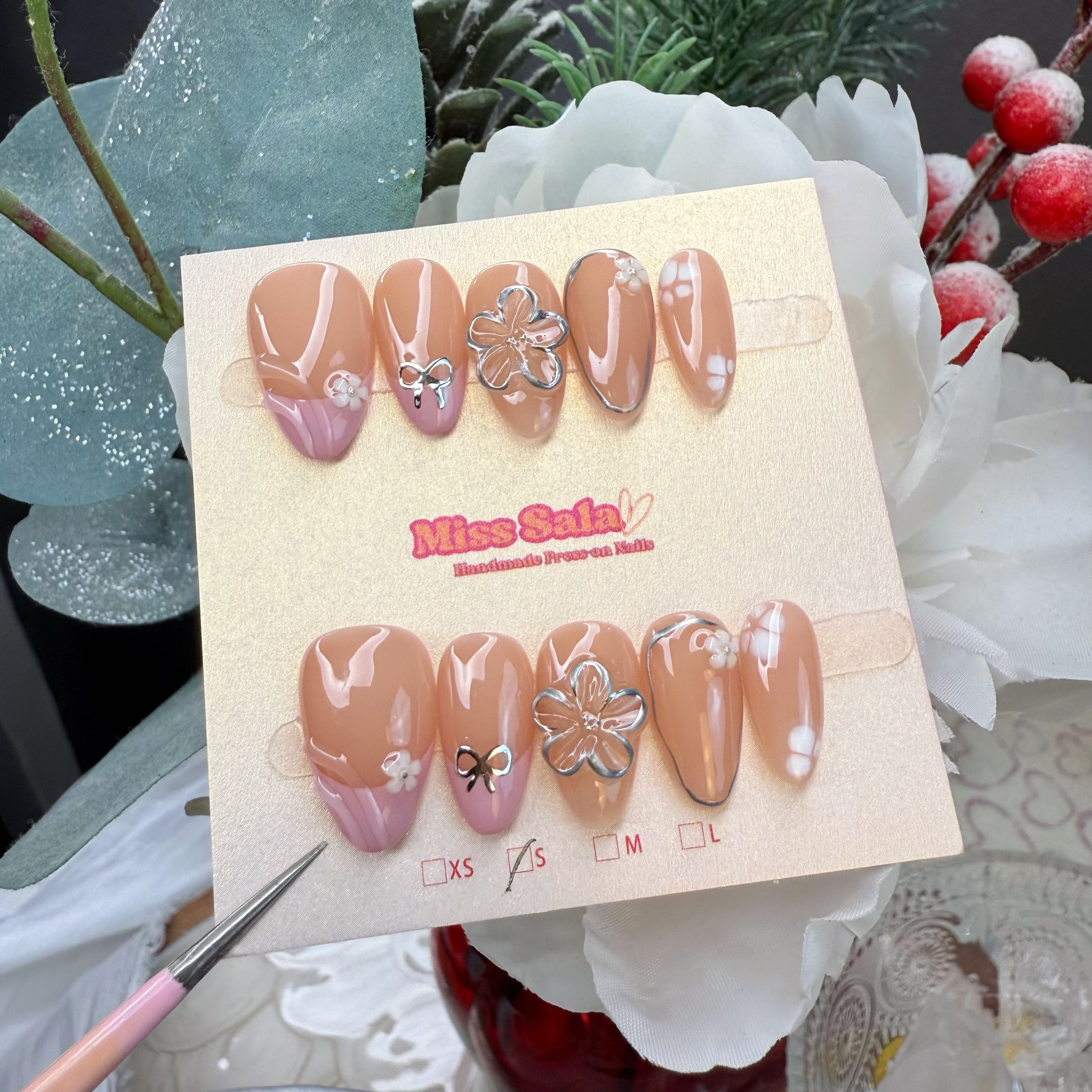 Almond-shaped press-on nails with a warm nude base, featuring solid pink and white curved French tips, 3D silver chrome flower and bow charms, and tiny white floral accents.