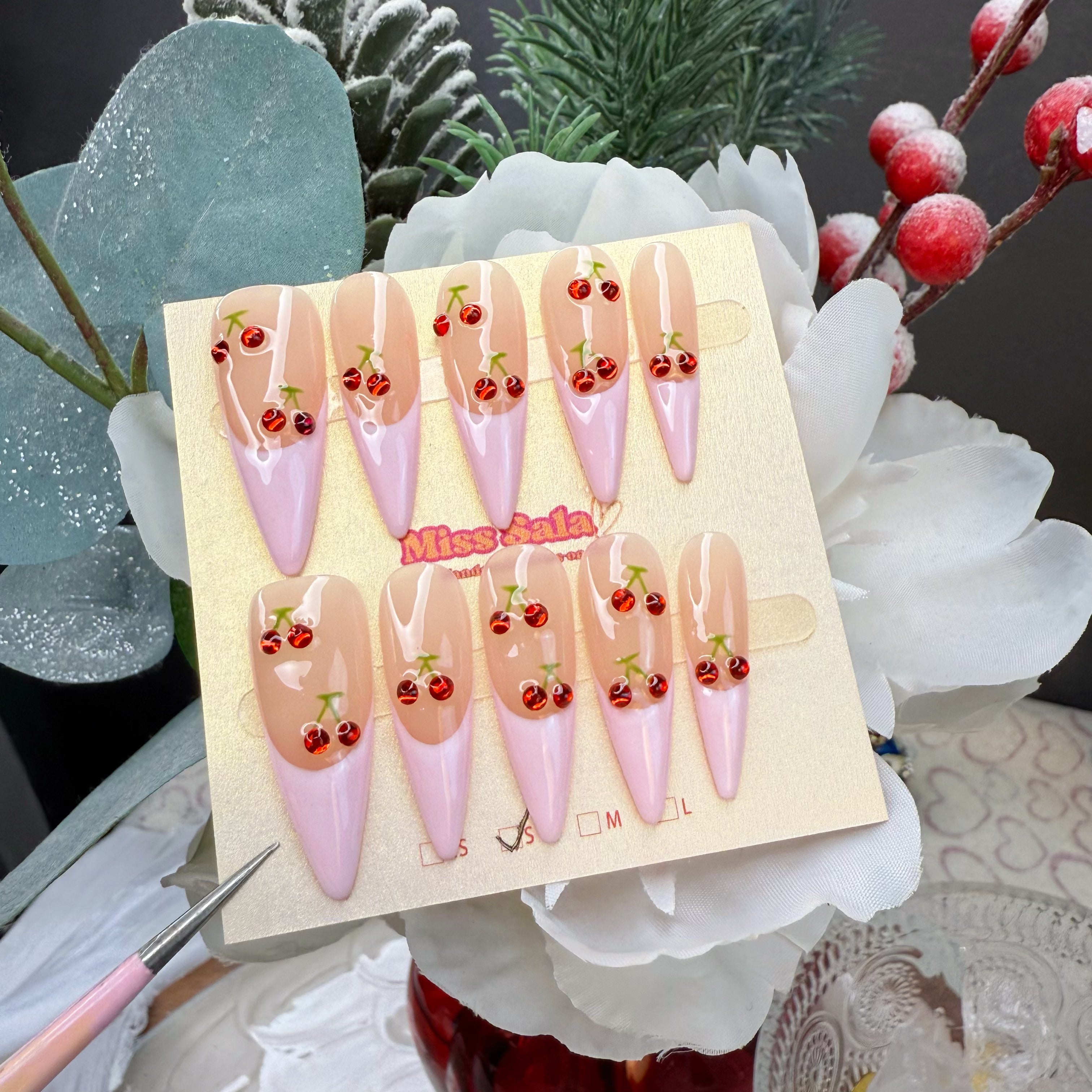 Long stiletto press-on nails with a glossy nude base, pastel pink French tips, and 3D red cherry charms with green stems.