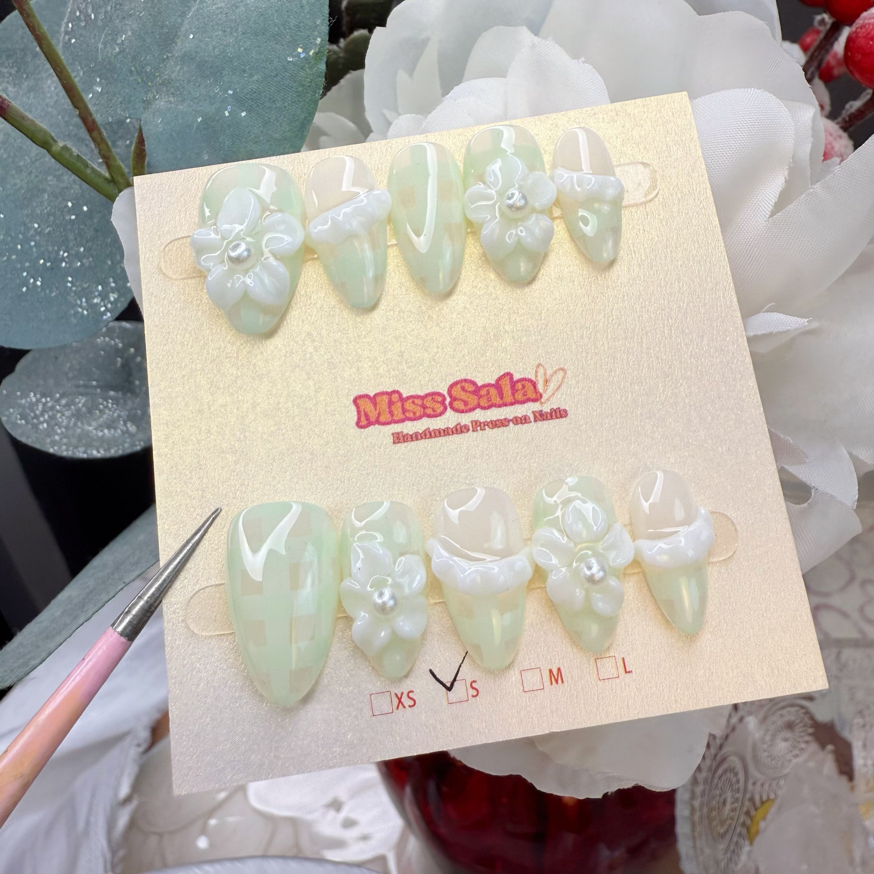 Almond press-on nails with a light green and cream plaid pattern, 3D white flower charms with pearl centers, and sculpted white ruffle accents.