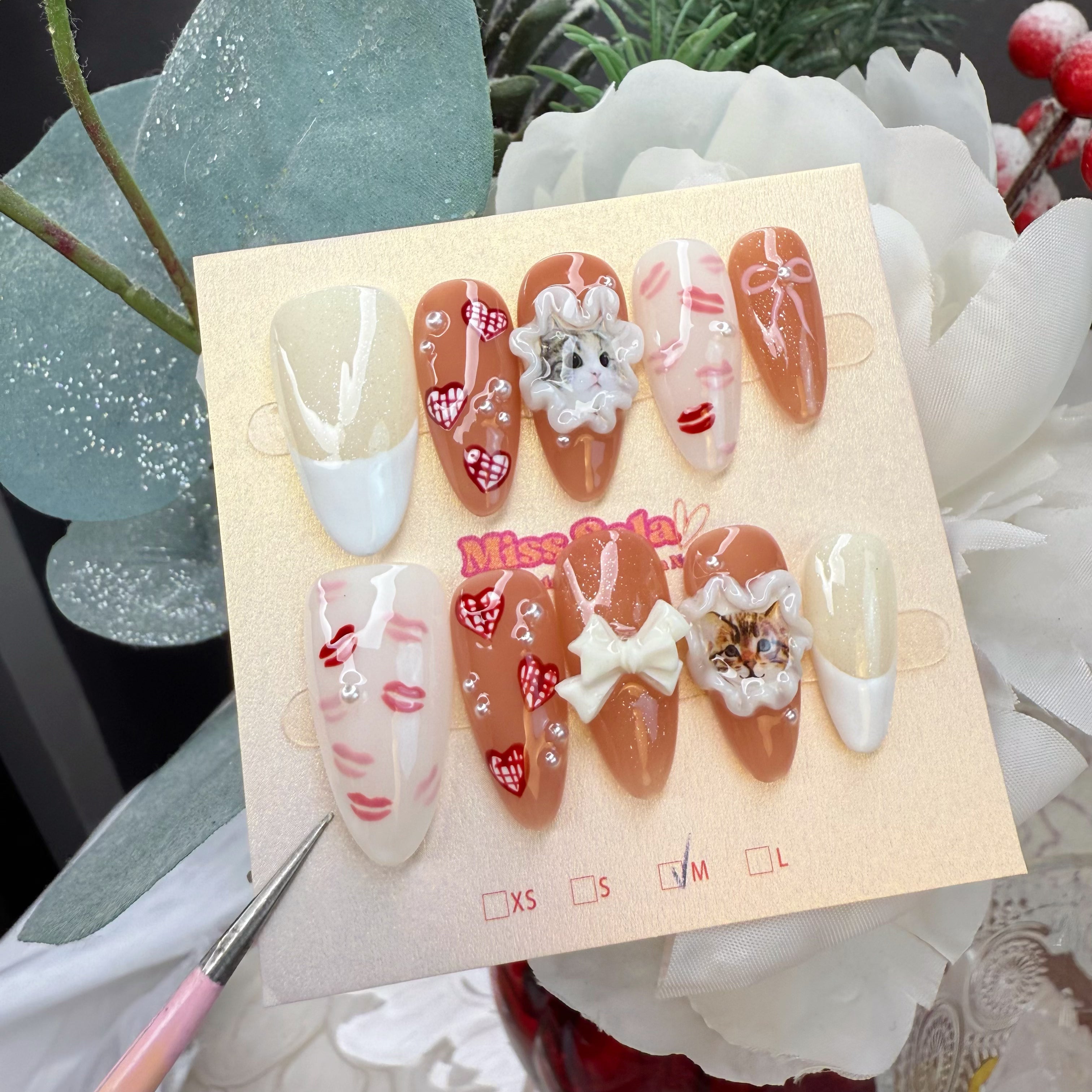 Almond-shaped press-on nails with a warm nude and white base, featuring 3D realistic cat portraits in ruffles, hand-painted red lip prints, red gingham hearts, 3D white bows, and pearls.