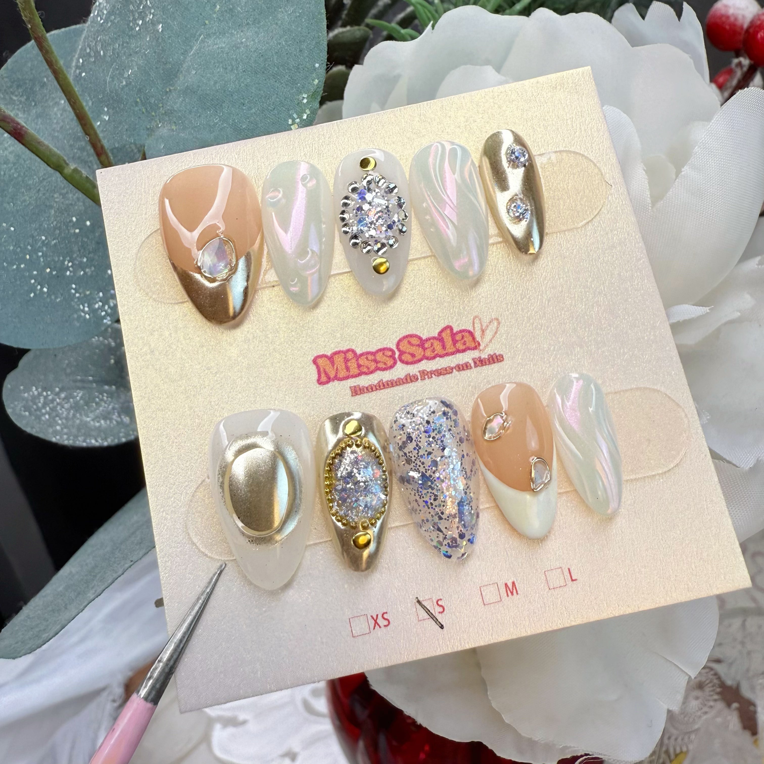 Almond-Shaped Press-On Nails with a Nude and White Base, Gold Chrome French Tips, Iridescent Pearl Finishes, and Ornate Crystal and Gold Stud Embellishments