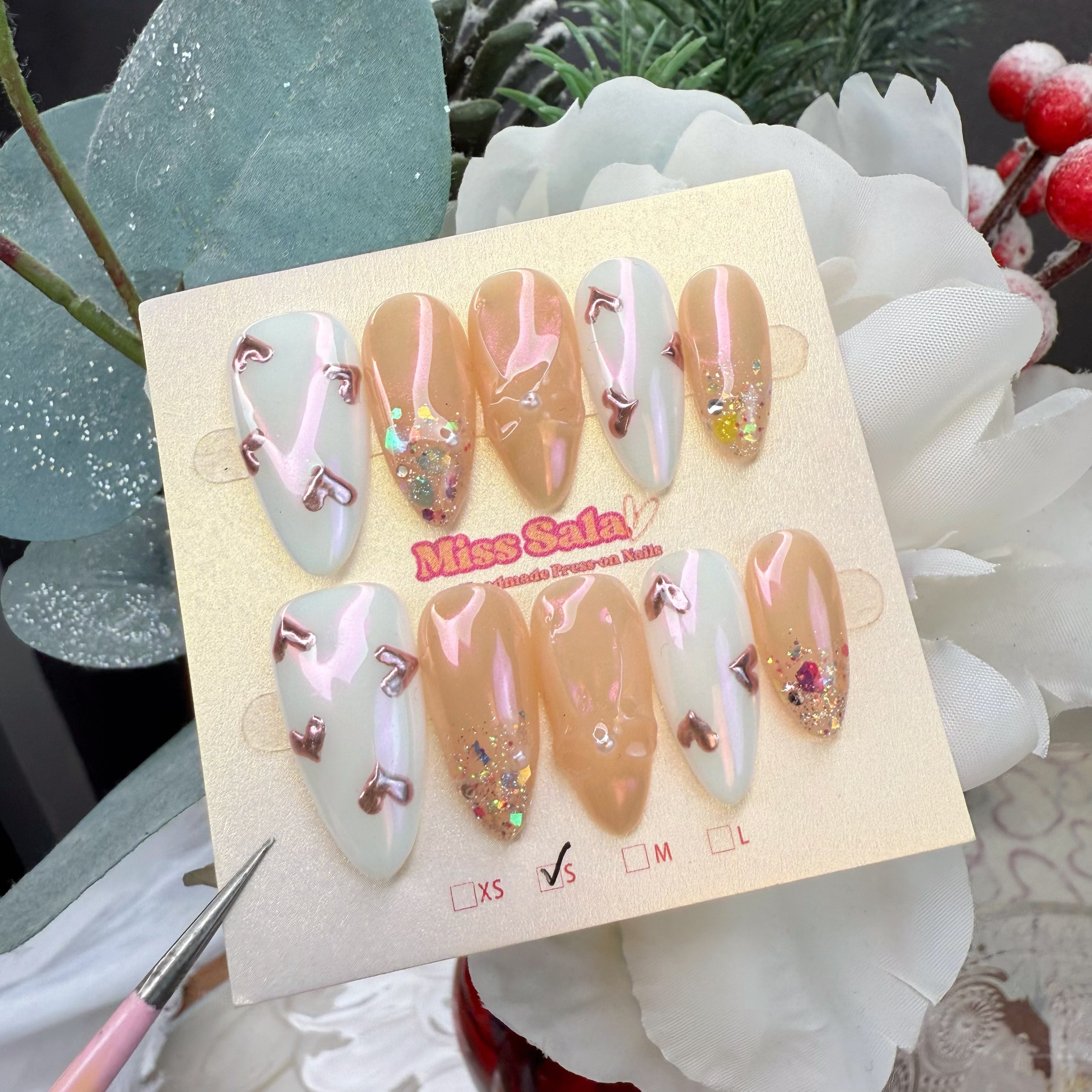 Almond-shaped press-on nails with an iridescent pearl and nude base, featuring rose gold metallic 3D hearts and chunky holographic glitter accents.