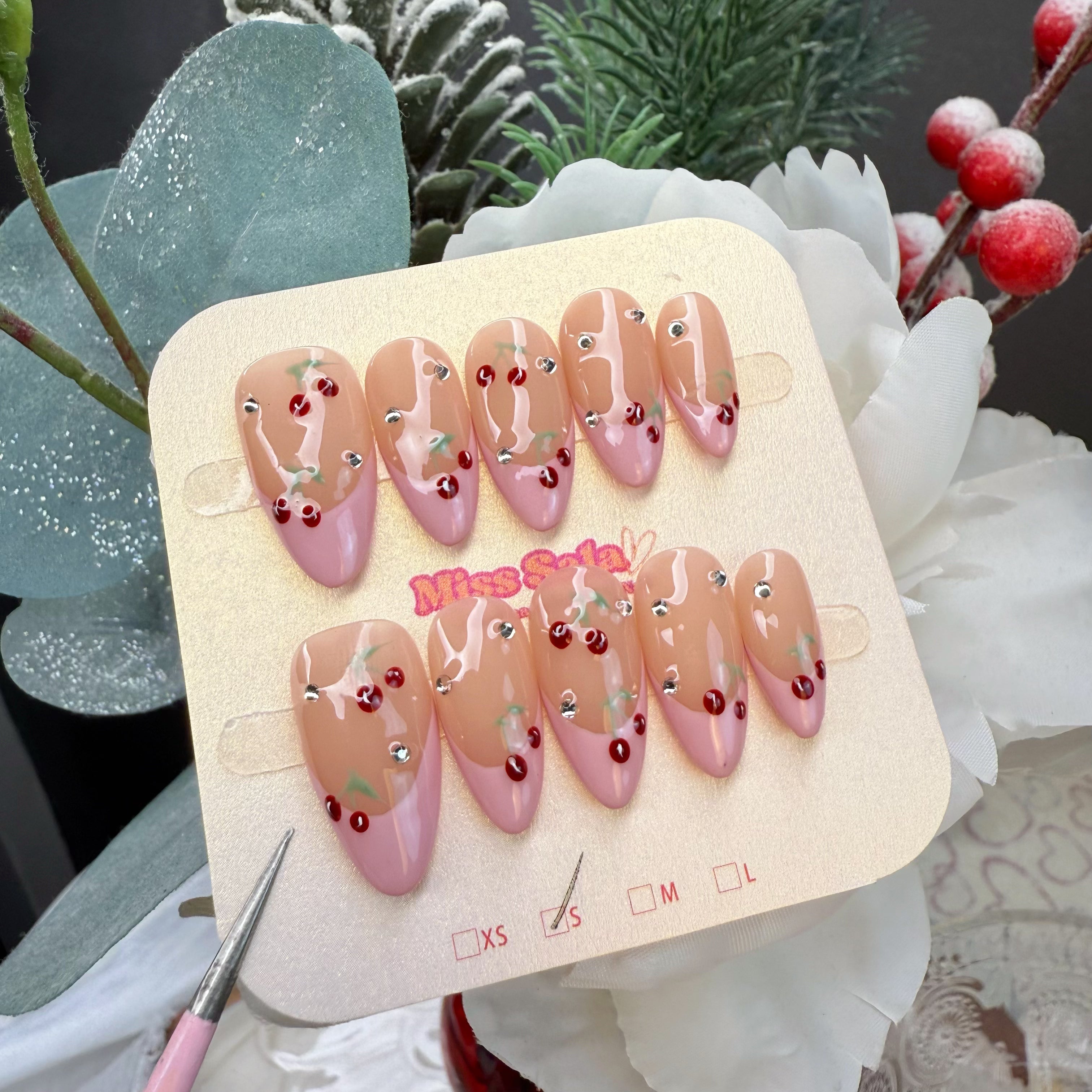 Almond-shaped press-on nails with a glossy nude base and soft pink French tips, featuring hand-painted red cherry motifs and sparkling rhinestone accents.