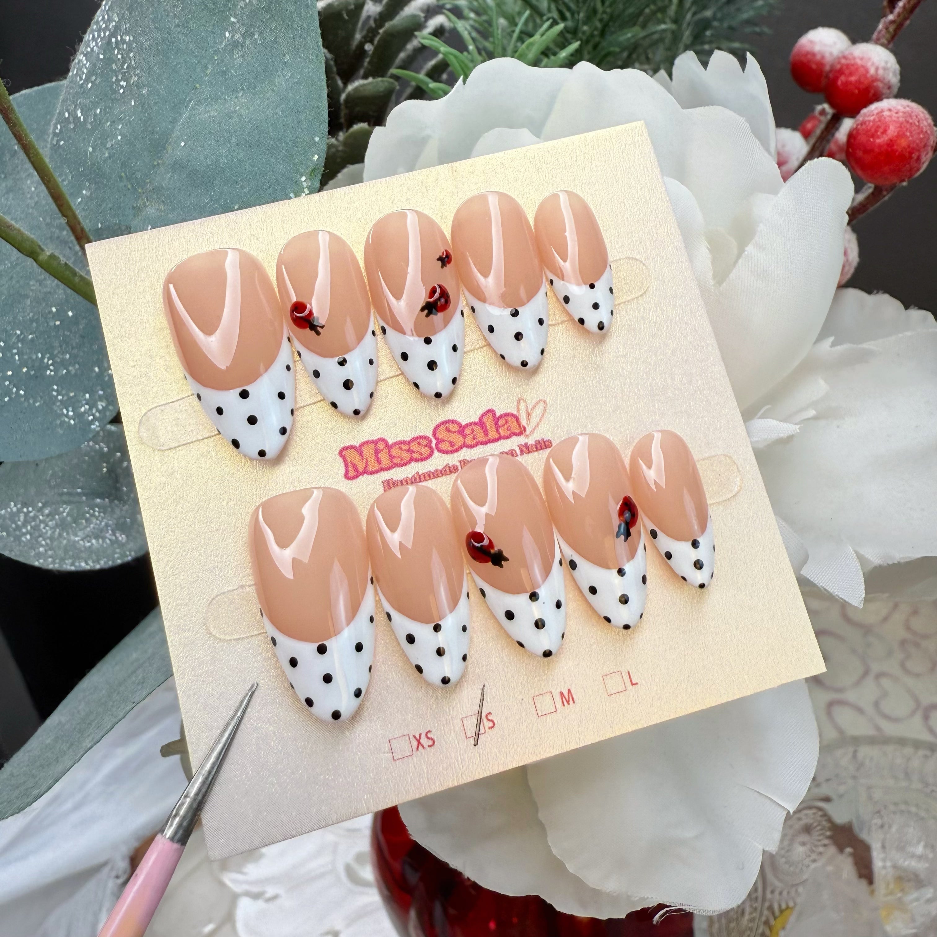 Almond press-on nails featuring a nude base with white polka dot French tips and hand-painted red ladybug accents.