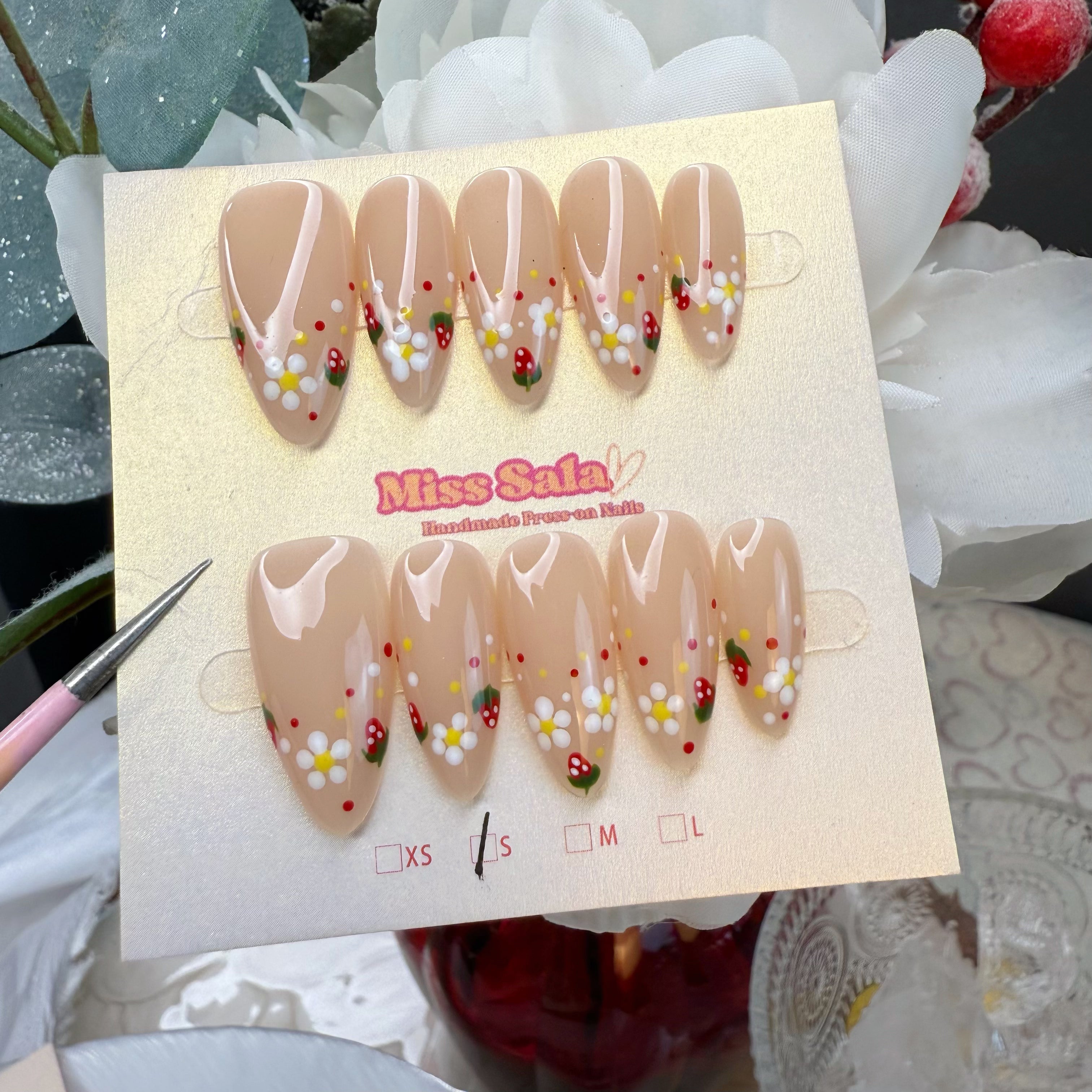 Almond-shaped press-on nails with a glossy nude base, featuring hand-painted white daisies with yellow centers and small red strawberries with green leaves.