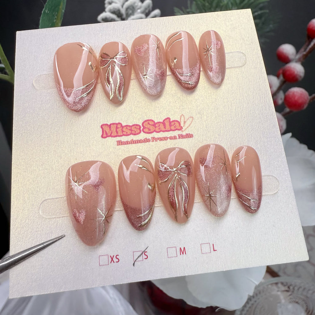 Almond-shaped press-on nails with a nude and shimmering rose-gold base, featuring hand-painted rose-gold glitter bows, metallic gold starbursts, and pink cat-eye glitter accents.